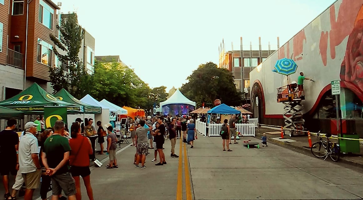 Crowd at a street art walk with tents and a mural being painted