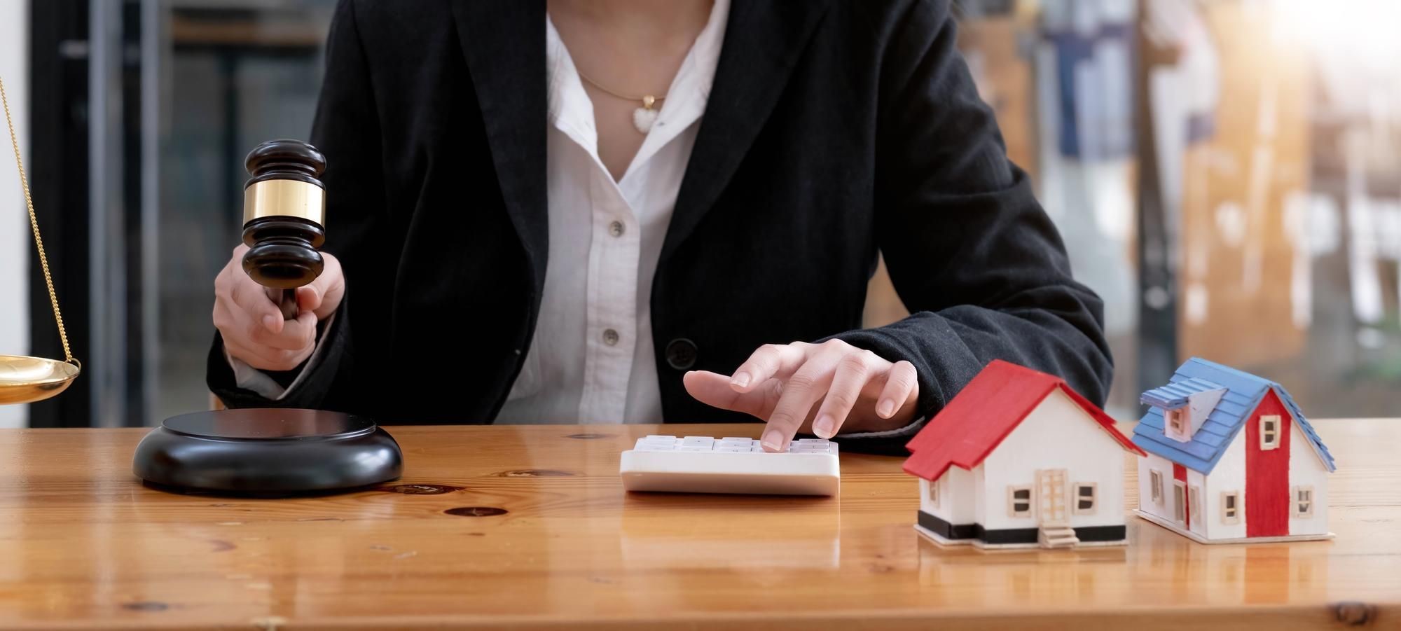 Person in a blazer holding a gavel beside a calculator and two toy houses