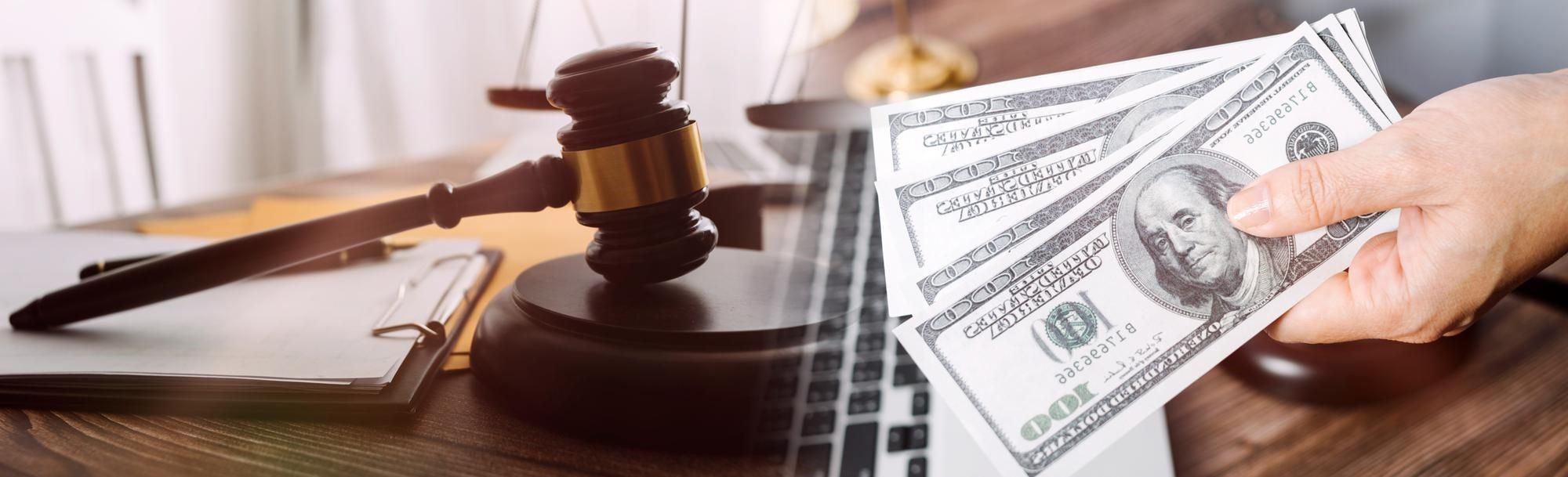 Gavel, scales, and a hand holding hundred-dollar bills on a desk