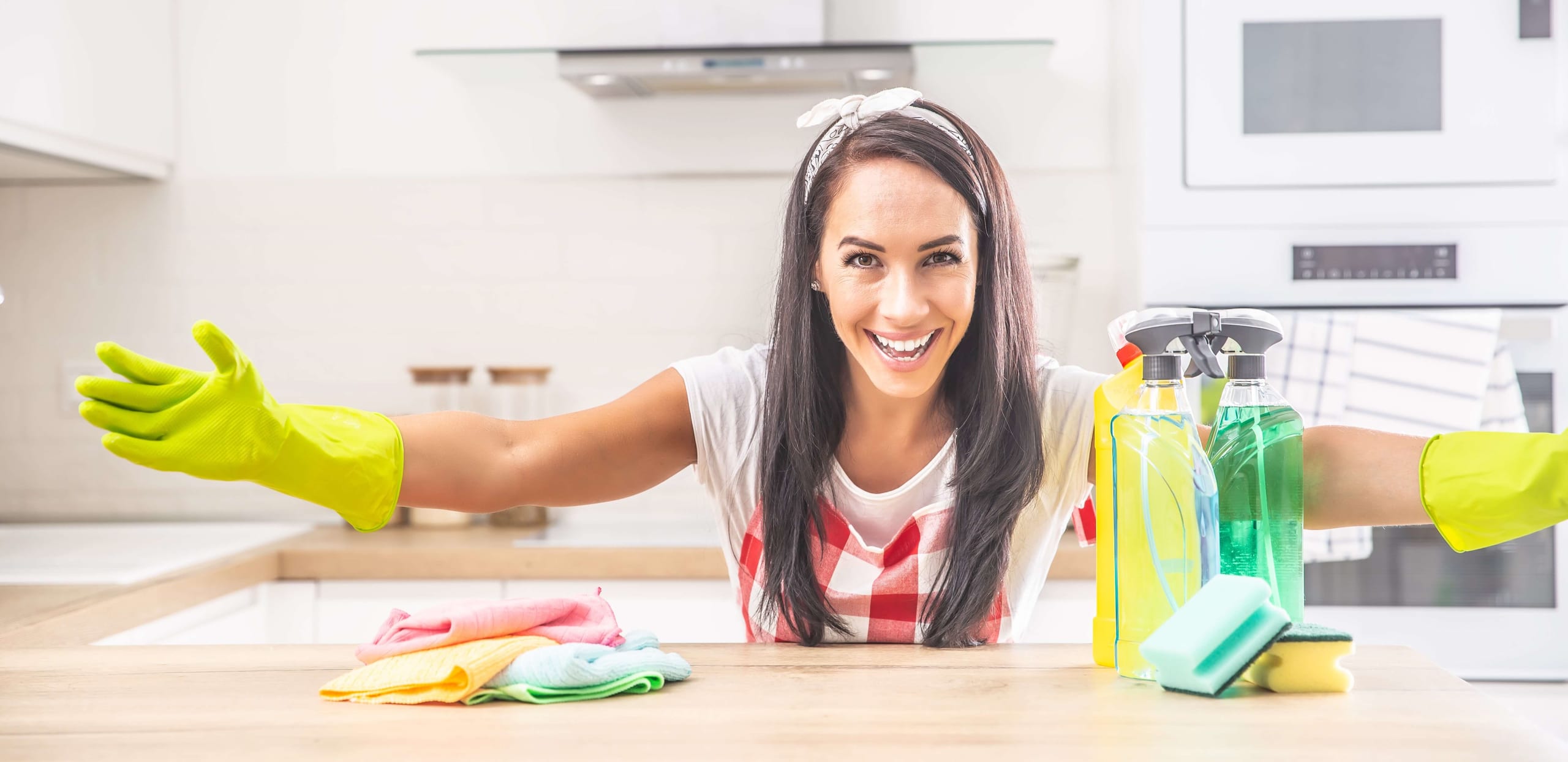 Woman in yellow cleaning gloves smiling at a kitchen island with spray bottles