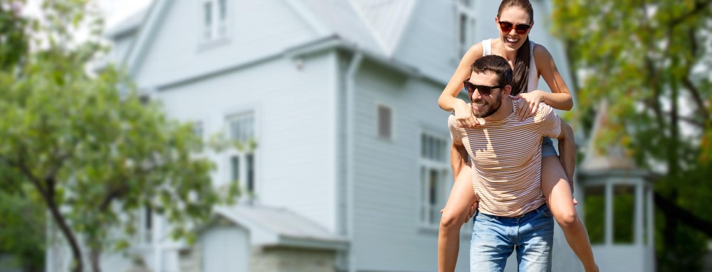 Smiling couple in sunglasses, the woman piggyback on the man, in front of a white house