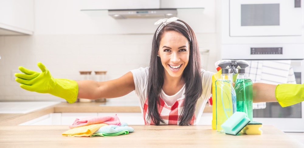 Smiling woman in yellow gloves with spray bottles on a kitchen island