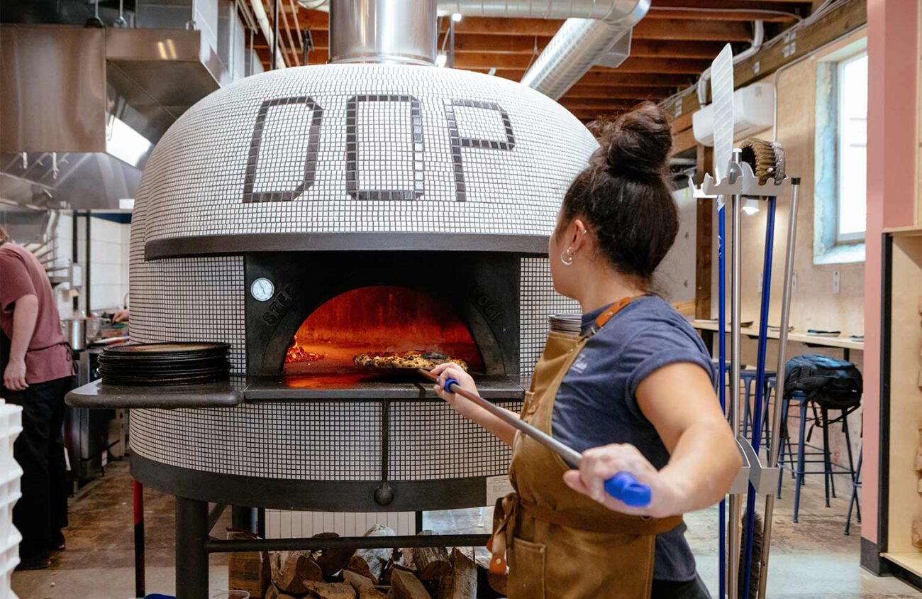 Cook sliding a pizza into a tiled DOP wood-fired oven