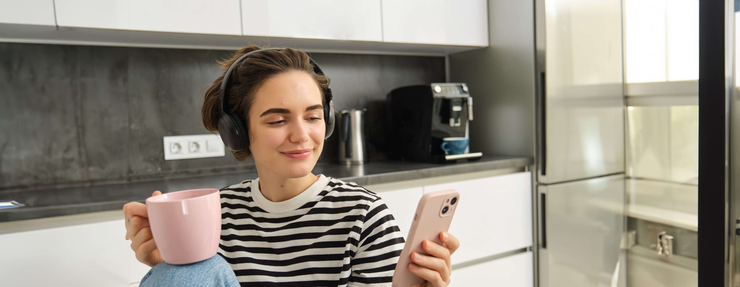Woman with headphones looking at a phone while holding a mug